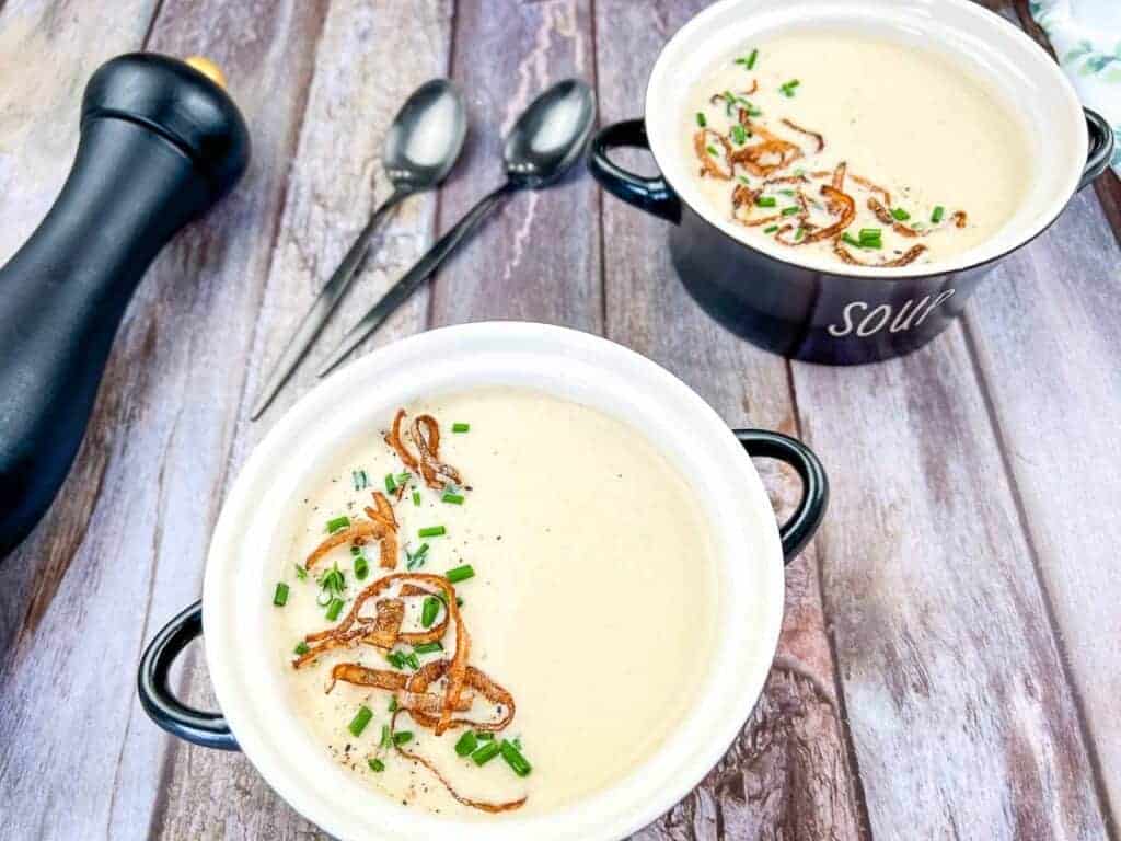 Two bowls of cream of onion soup topped with crispy onions and chives on a wooden table.