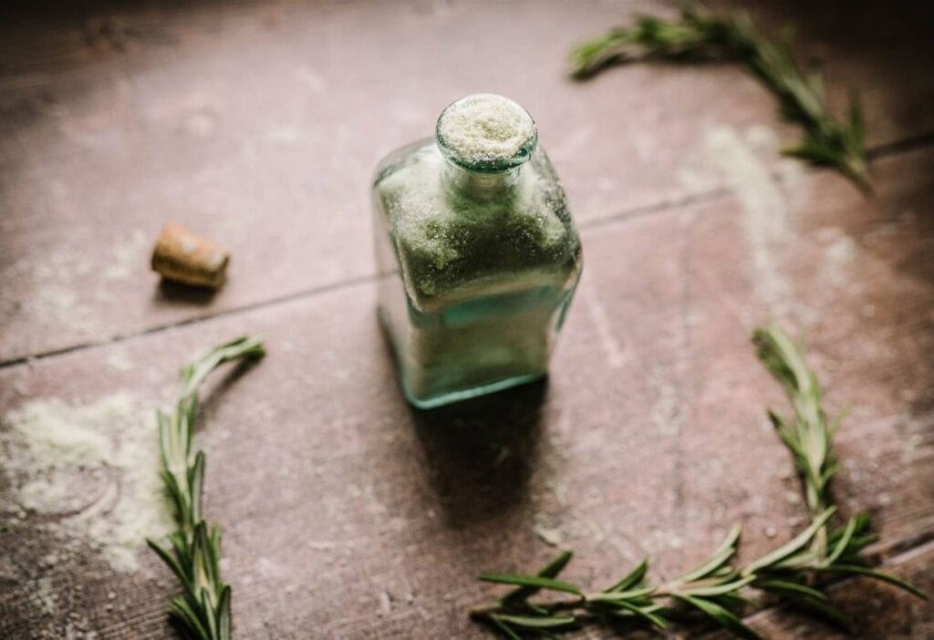 A square glass bottle filled with coarse salt on a wooden surface, surrounded by rosemary sprigs and a cork nearby.