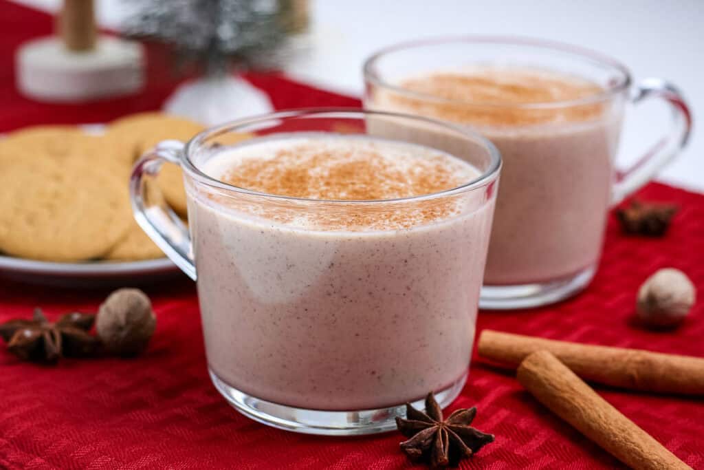 Two clear mugs filled with creamy spiced beverage, topped with cinnamon, on a red cloth. Nearby are cookies, cinnamon sticks, star anise, and nutmeg.