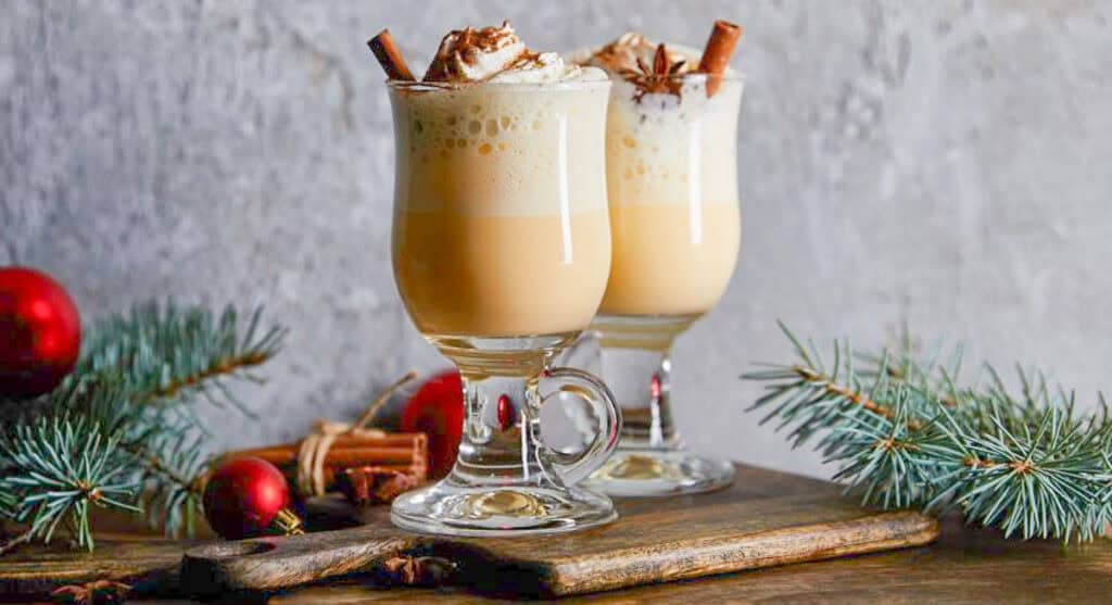 Two glass mugs of creamy eggnog topped with whipped cream and cinnamon sticks on a wooden board. Pine branches and red ornaments decorate the scene.