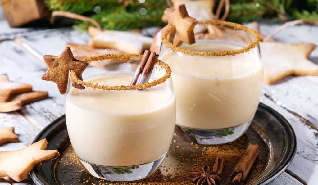 Two glasses of eggnog garnished with cinnamon sticks and star-shaped cookies on a tray, surrounded by more cookies and greenery in the background.