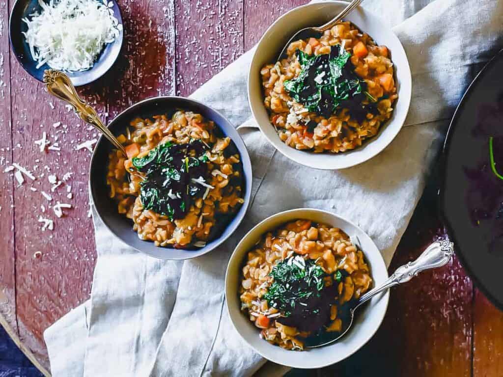 Three bowls of risotto topped with chopped greens sit on a cloth on a wooden table, with a dish of grated cheese beside them.