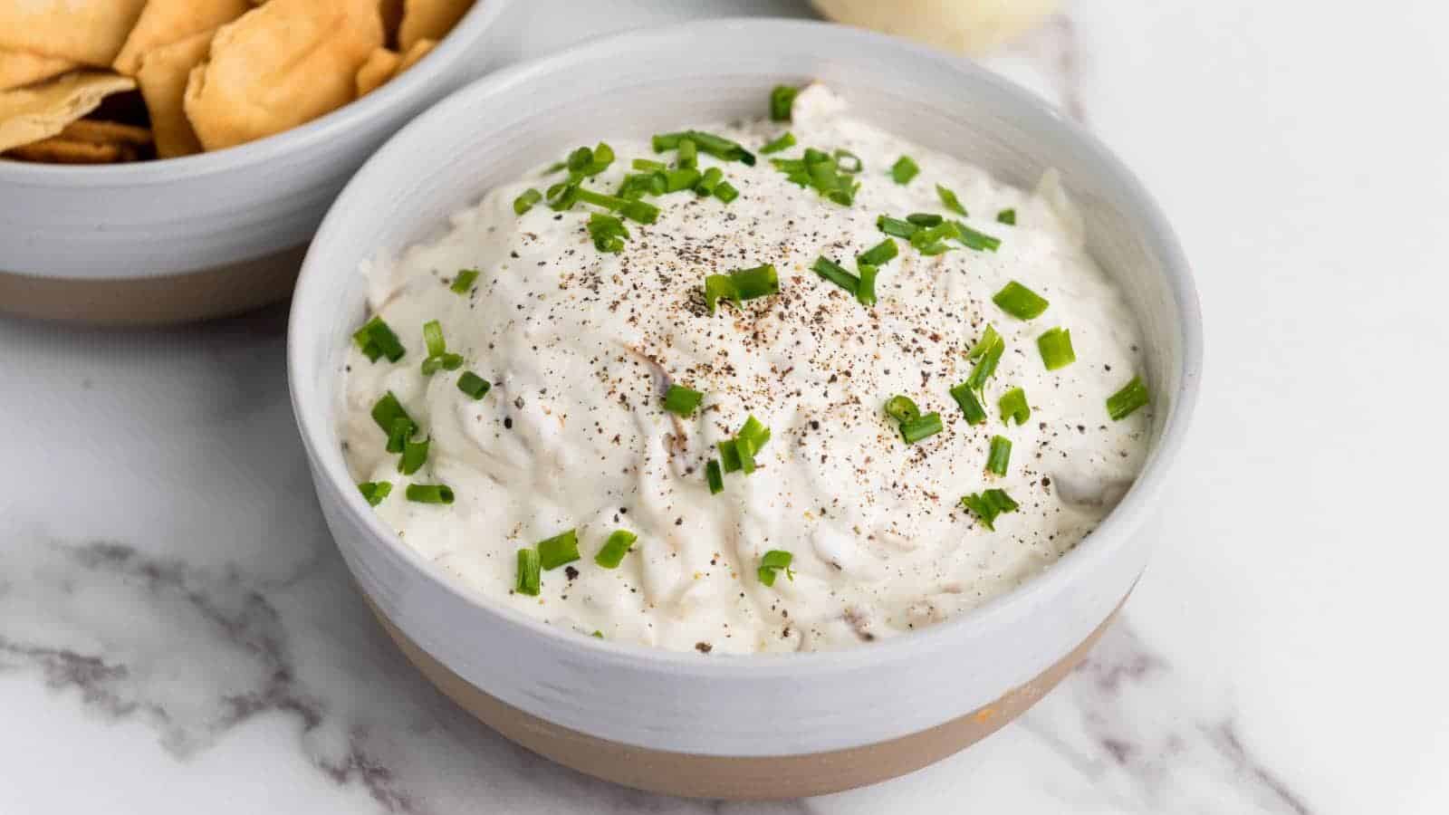 A bowl of creamy french onion dip topped with chopped chives and black pepper, placed beside a bowl of chips on a marble surface.