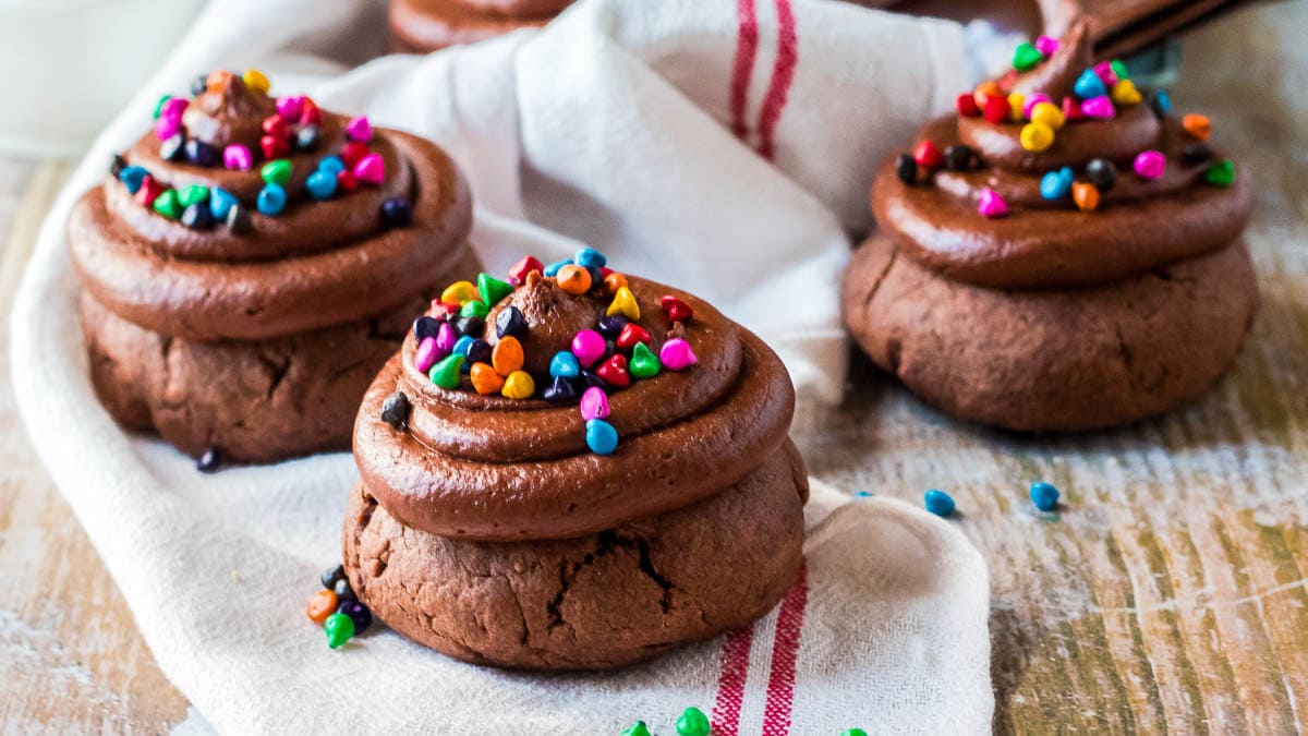 Chocolate cookies topped with chocolate frosting and colorful sprinkles on a white cloth.