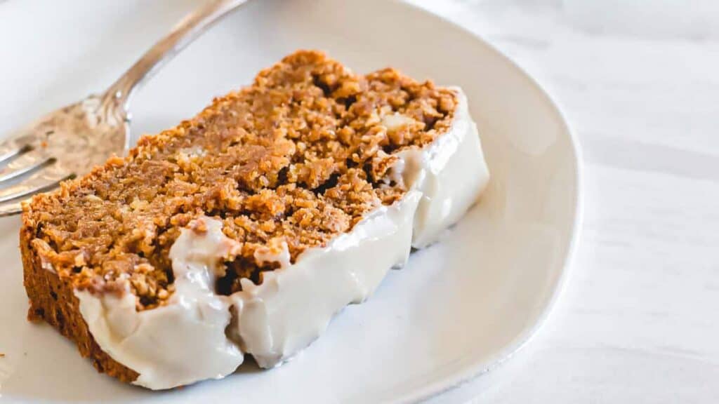 A slice of banana bread with white icing on a plate, accompanied by a fork.