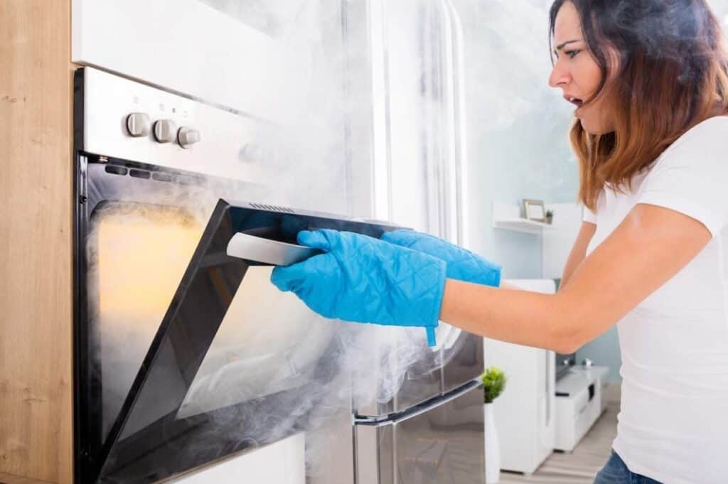 A woman in a white shirt and blue oven mitts opens a smoking oven in a kitchen, expressing concern.