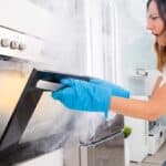 A woman in a white shirt and blue oven mitts opens a smoking oven in a kitchen, expressing concern.