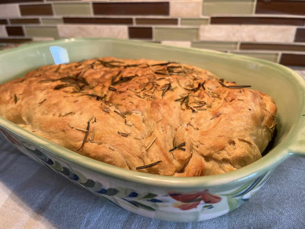 A rosemary-topped loaf of bread in a decorative baking dish, placed on a tablecloth with a tiled backsplash in the background.