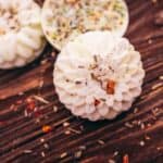 A round, intricately patterned soap with dried herbs rests on a wooden surface, surrounded by scattered herb pieces.