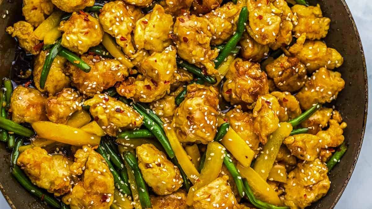 A dish with fried chicken pieces, yellow bell peppers, and green beans, topped with sesame seeds, in a pan.