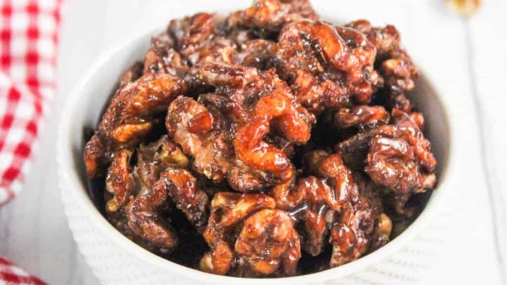 A white bowl filled with glazed candied walnuts on a white surface, next to a red and white checkered cloth.