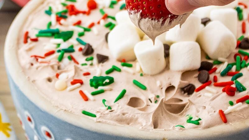 A hand dips a strawberry into a bowl of whipped dip topped with marshmallows and sprinkles. 