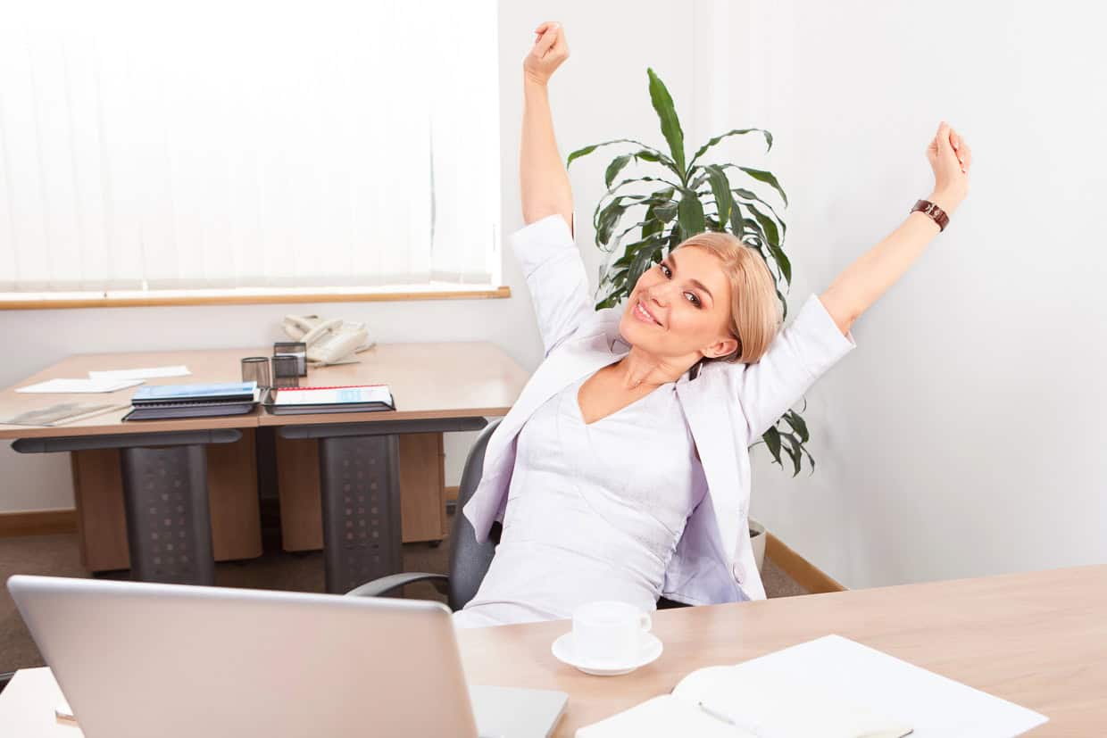 Person in a light-colored outfit stretching at a desk with a laptop and a cup of coffee, in a bright office setting.