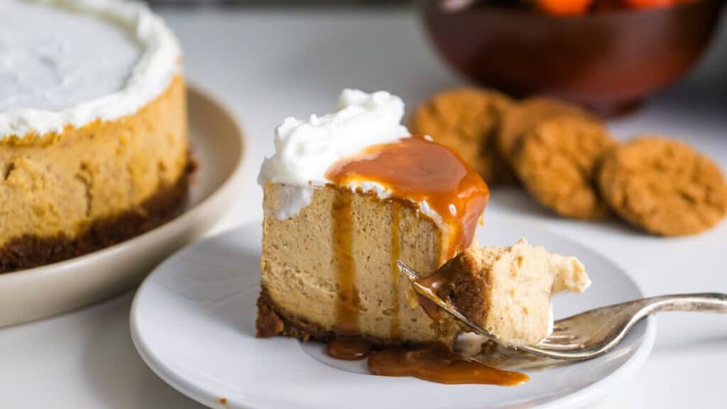 A slice of cheesecake topped with caramel sauce and whipped cream on a white plate. A fork holds a bite. Whole cheesecake and cookies are in the background.
