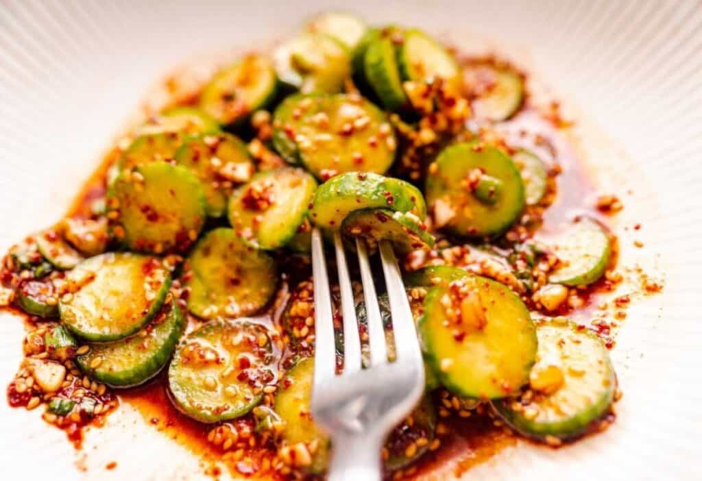 A dish of sliced cucumbers is mixed with a spicy red sauce, garnished with sesame seeds and garlic. A fork rests on the edge of the plate.