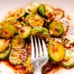 A dish of sliced cucumbers is mixed with a spicy red sauce, garnished with sesame seeds and garlic. A fork rests on the edge of the plate.