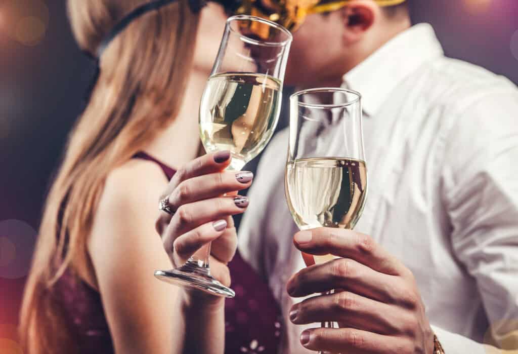 Two people clinking glasses of champagne, both wearing masks, with festive background lighting.