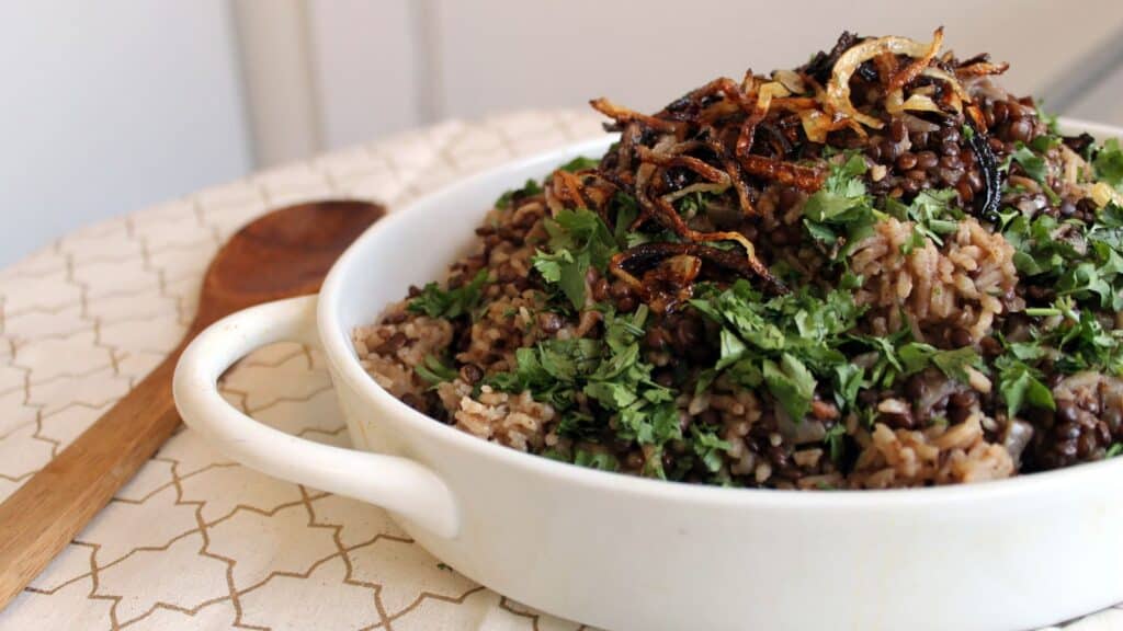 A white dish filled with lentils, rice, herbs, and topped with caramelized onions. A wooden spoon is nearby.