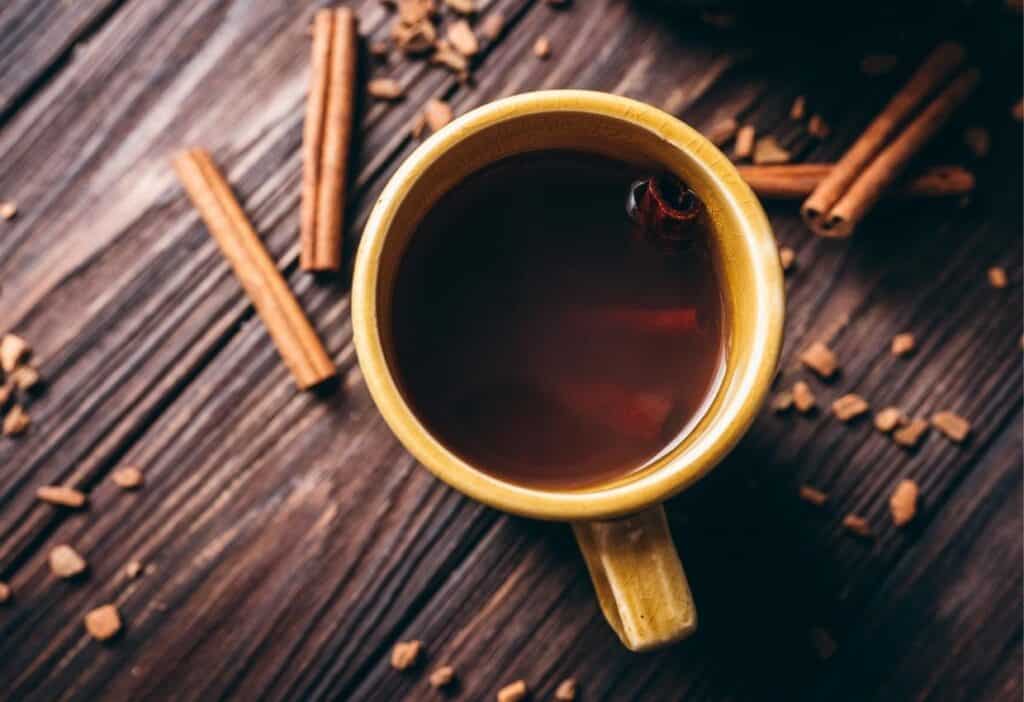 A yellow mug filled with dark liquid on a wooden surface, surrounded by cinnamon sticks and small scattered pieces.