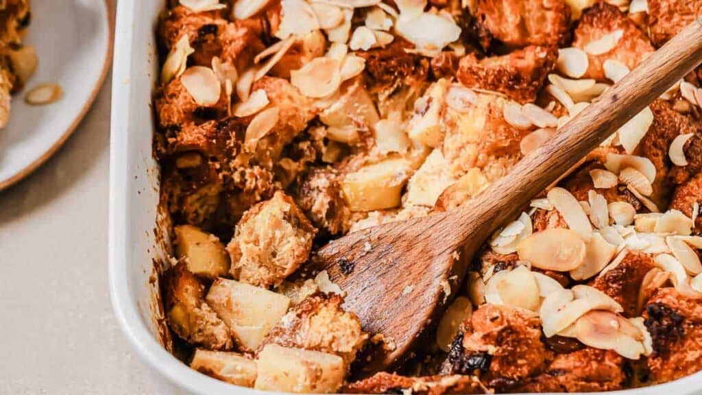 Close-up of a baked bread pudding with visible sliced almonds and chunks of apple in a casserole dish, with a wooden spoon partially submerged in the dish.