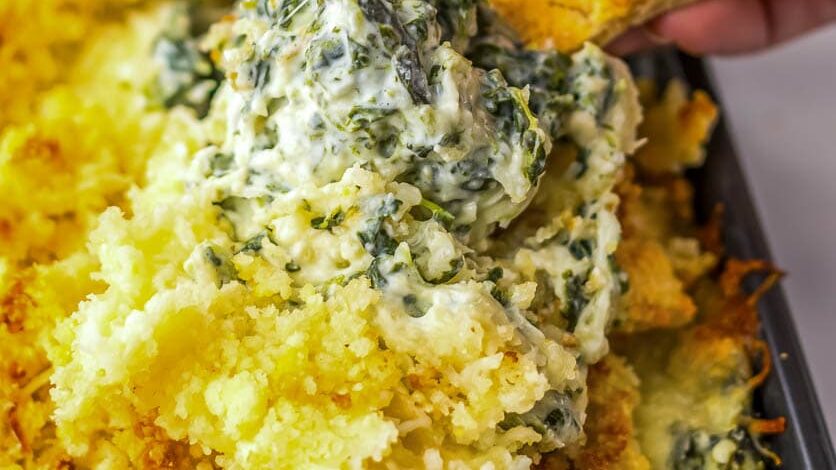 A hand dips a chip into a creamy spinach artichoke dip topped with breadcrumbs.