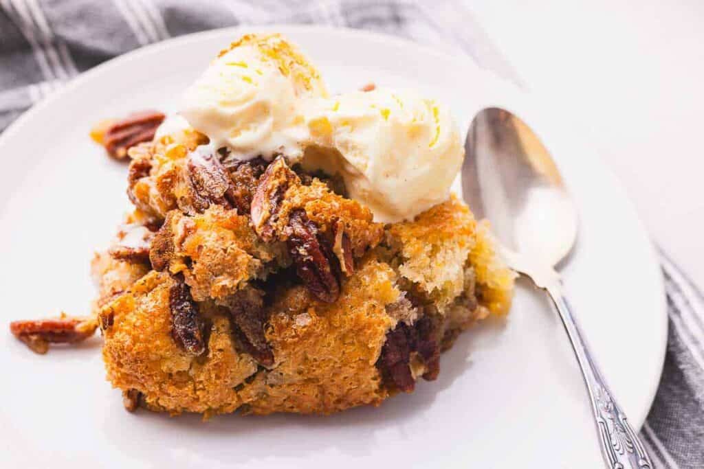 A slice of pecan pie topped with two scoops of vanilla ice cream on a white plate with a spoon.