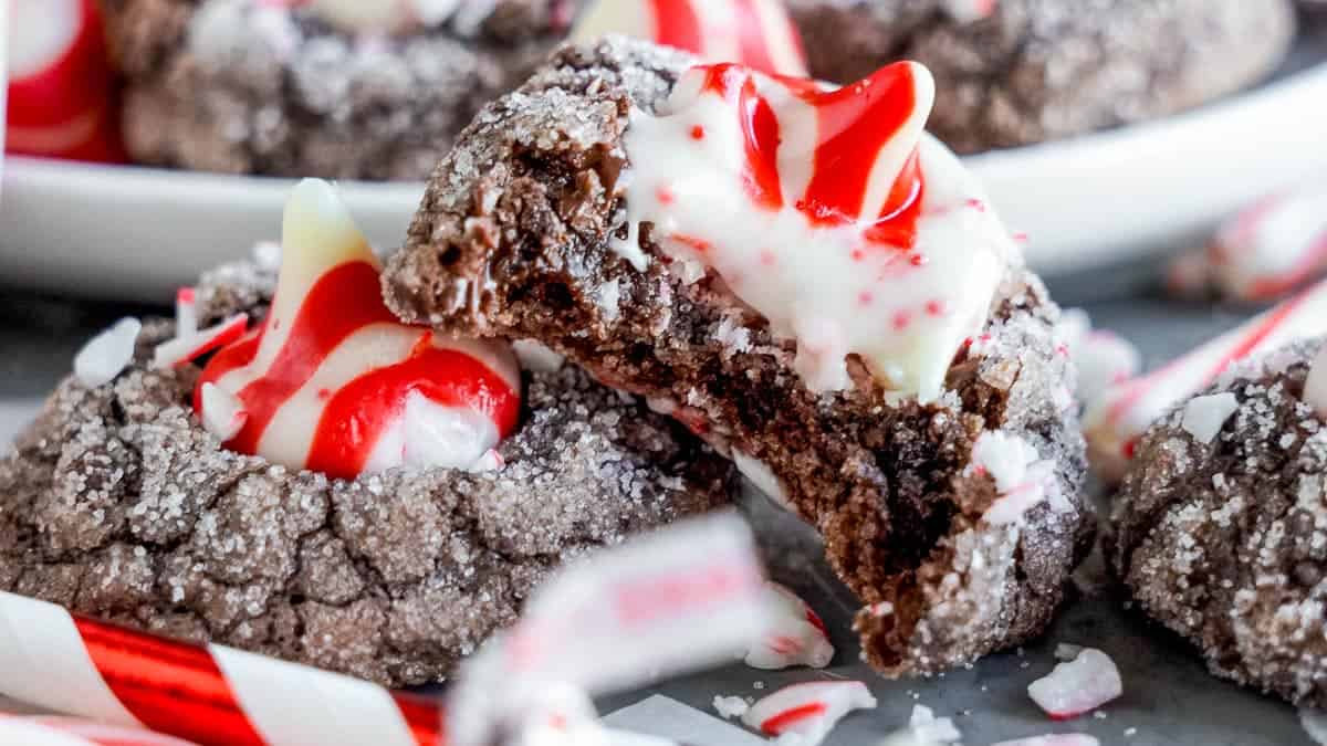 Chocolate cookies topped with red and white peppermint swirls and sprinkled with sugar, with one cookie partially eaten, surrounded by broken candy canes.