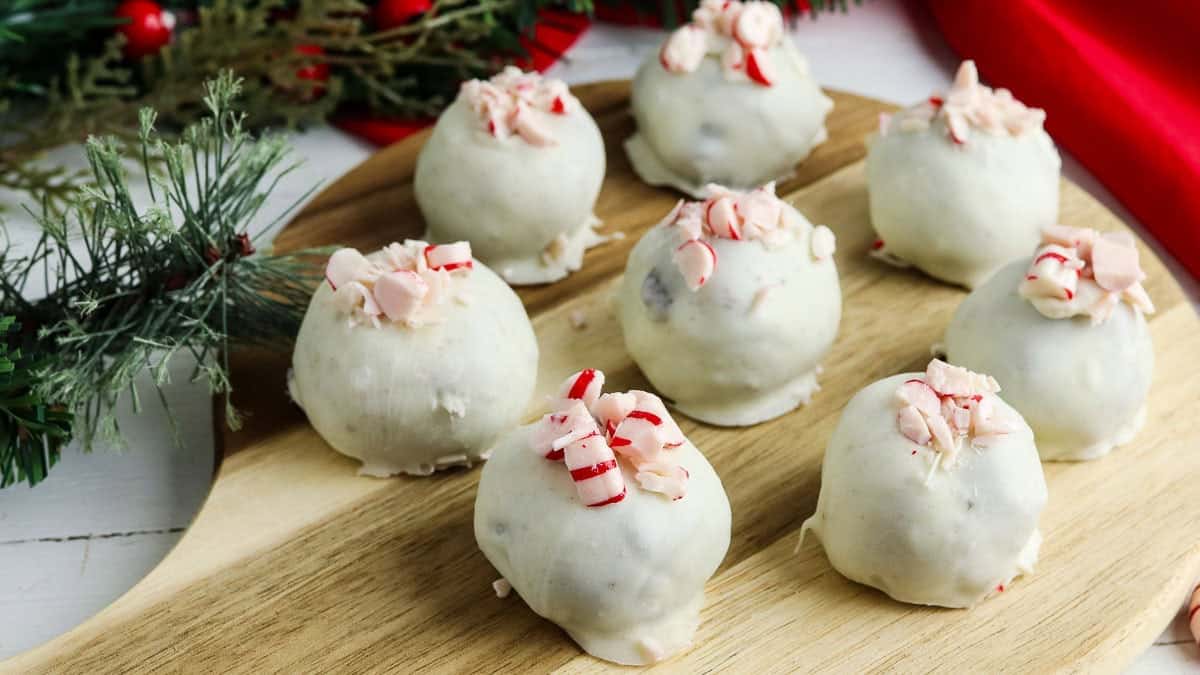 A wooden board displays eight white chocolate-covered balls topped with crushed peppermint, surrounded by holiday greenery, red fabric, Oreo cookies, and peppermint candies.