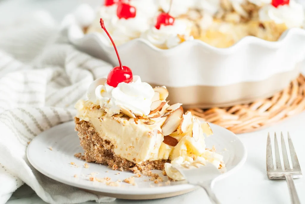 A slice of creamy dessert topped with whipped cream, a cherry, and almond slices on a plate, with a fork beside it. Background shows the remaining dessert in a dish.