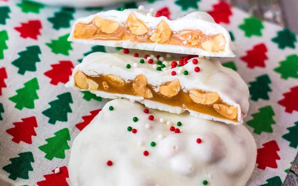 White chocolate-covered candies with peanuts and caramel filling, topped with red and green sprinkles, on a festive Christmas tree-patterned napkin.