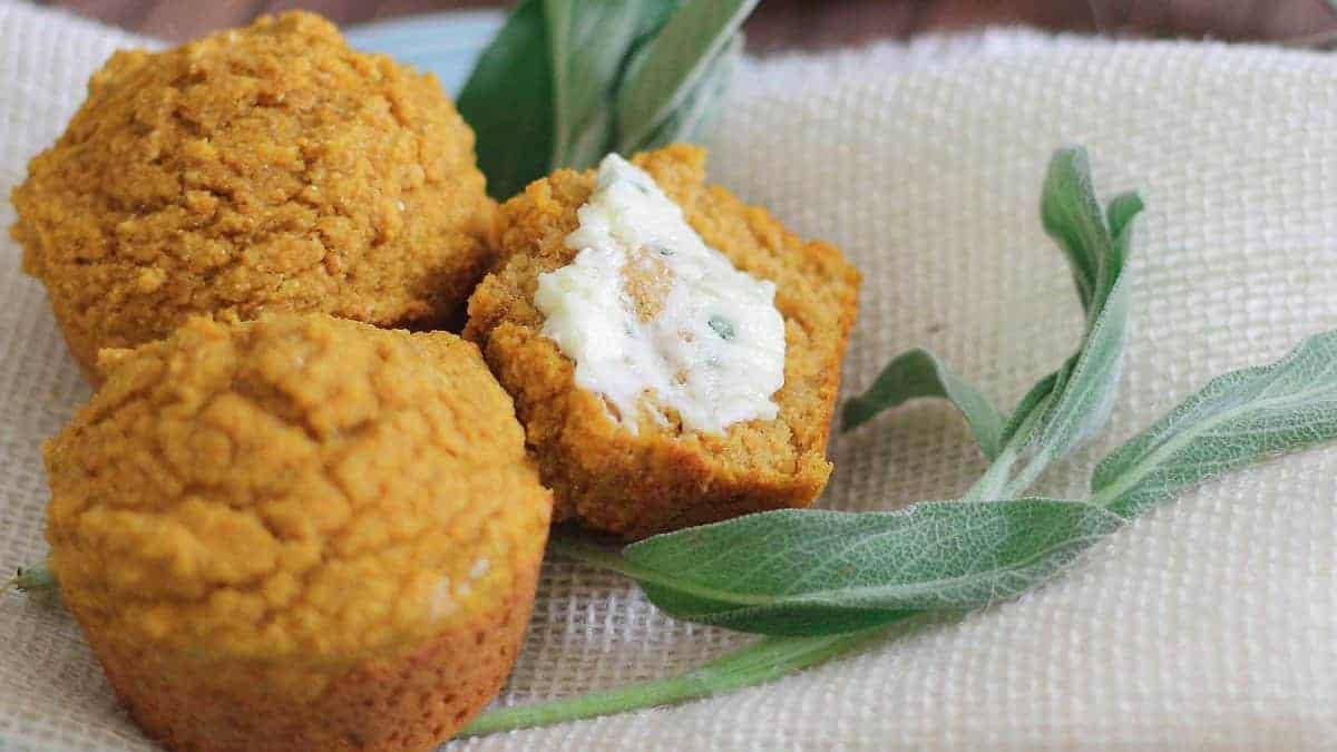 Three pumpkin muffins on burlap with a sprig of sage. One muffin is halved and spread with butter.