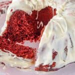 A ring-shaped red velvet cake with white icing is sliced to reveal its red interior, displayed on a pink cake stand.