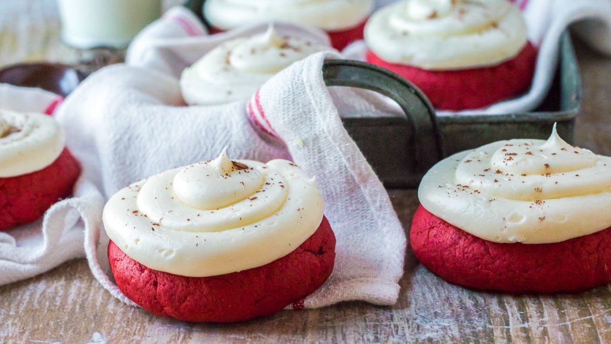 Red velvet cookies topped with white frosting are arranged on a wooden table. A metal tray and cloth are visible in the background.