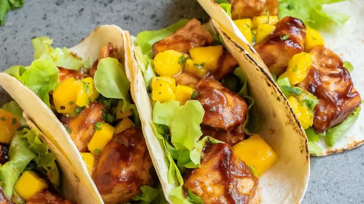 Three chicken tacos with lettuce and mango salsa are arranged on a gray surface. There is a small green lime and a bowl of mango salsa on the side.