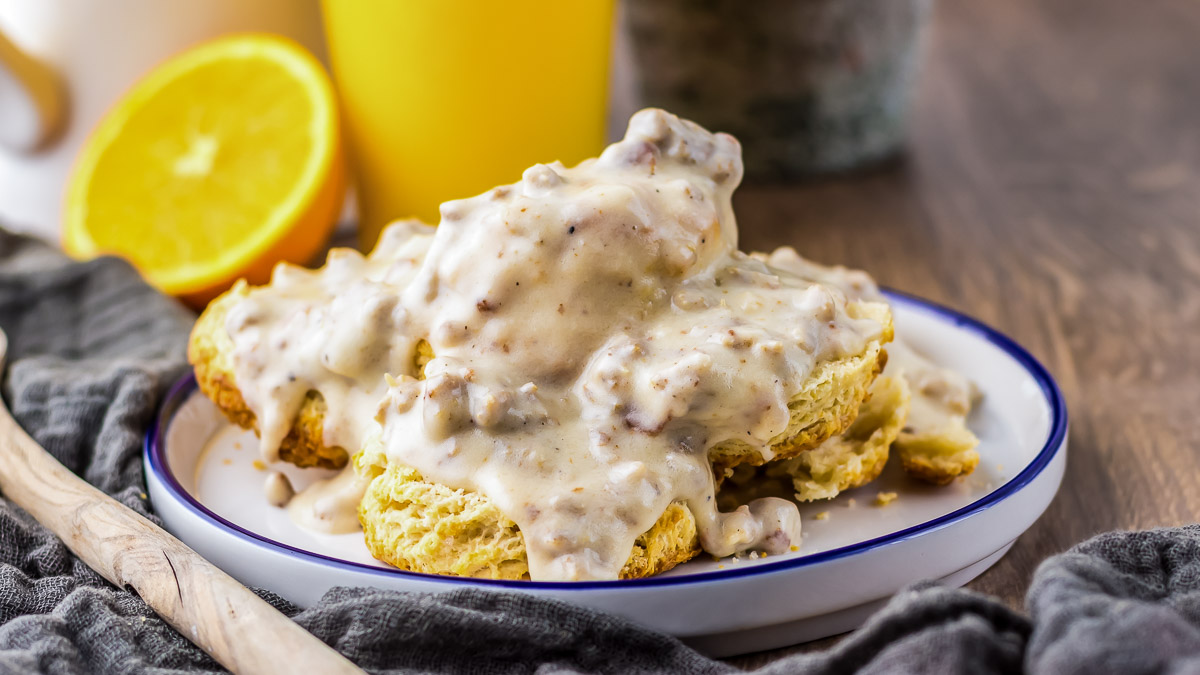 A plate of biscuits covered in creamy sausage gravy sits on a table, accompanied by a halved orange and a yellow mug in the background.