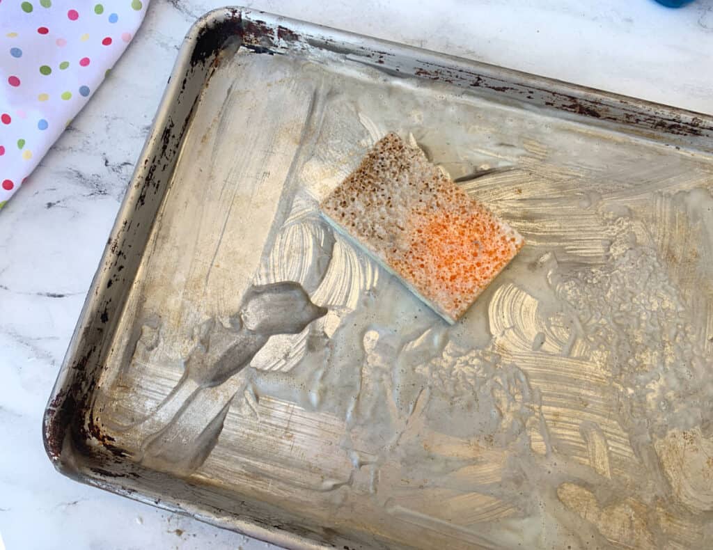 Dirty baking sheet with used sponge on top, featuring grease marks and food residue.