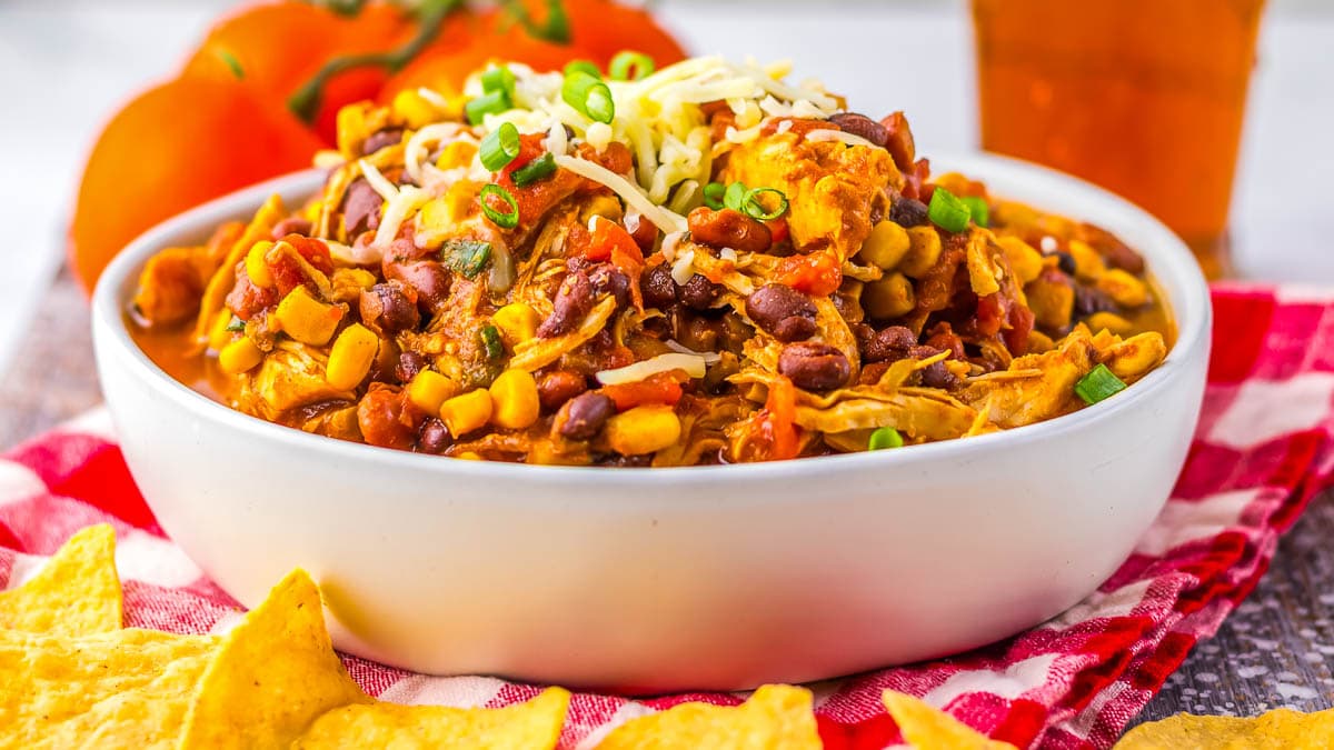 A bowl of chicken chili topped with cheese and green onions, surrounded by tortilla chips.