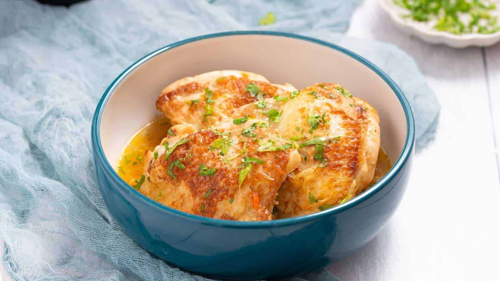 A blue bowl of cooked chicken breasts garnished with chopped herbs on a light fabric backdrop.