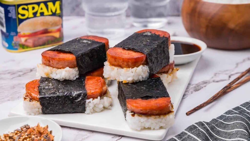 Four pieces of Spam musubi on a rectangular white plate with a small dish of soy sauce and a can of Spam in the background.