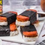 Four pieces of Spam musubi on a rectangular white plate with a small dish of soy sauce and a can of Spam in the background.