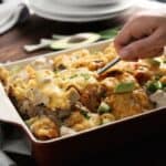 A hand using a fork to serve a portion of casserole topped with melted cheese and garnished with avocado in a baking dish, perfect for making comfort food without all the calories.