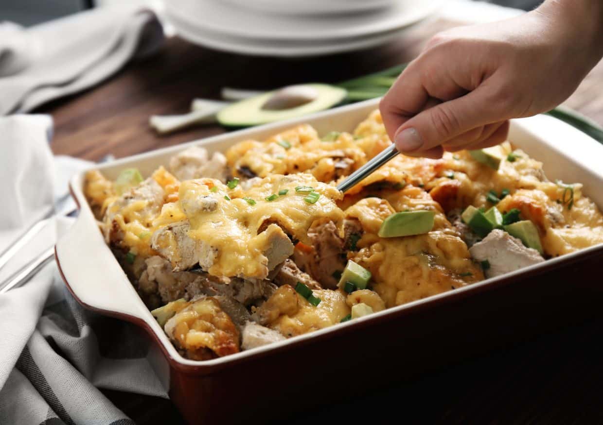 A hand using a fork to serve a portion of casserole topped with melted cheese and garnished with avocado in a baking dish, perfect for making comfort food without all the calories.