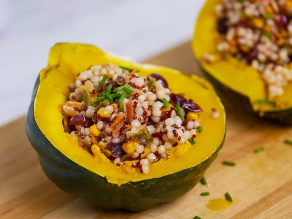 Halved acorn squash filled with couscous, quinoa, nuts, and vegetables on a wooden board.