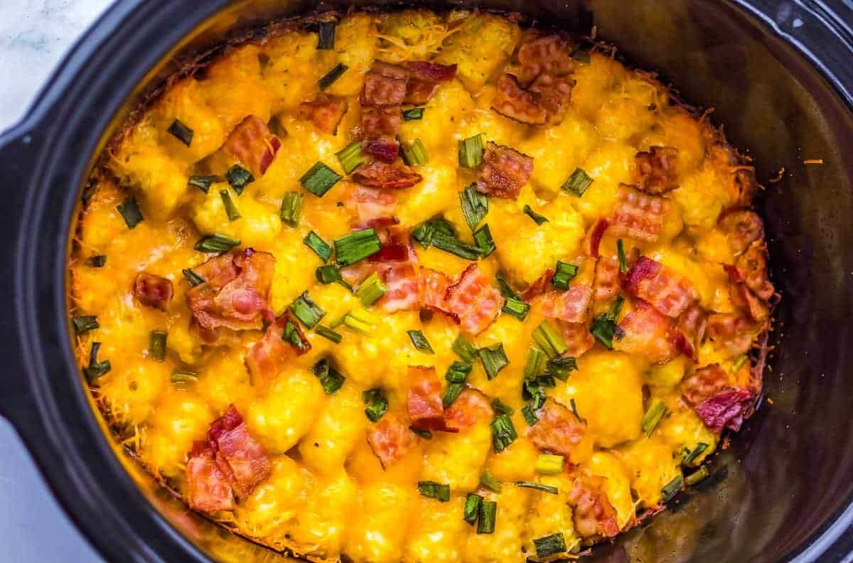 A crockpot filled with tater tots, melted cheese, bacon pieces, and chopped green onions.