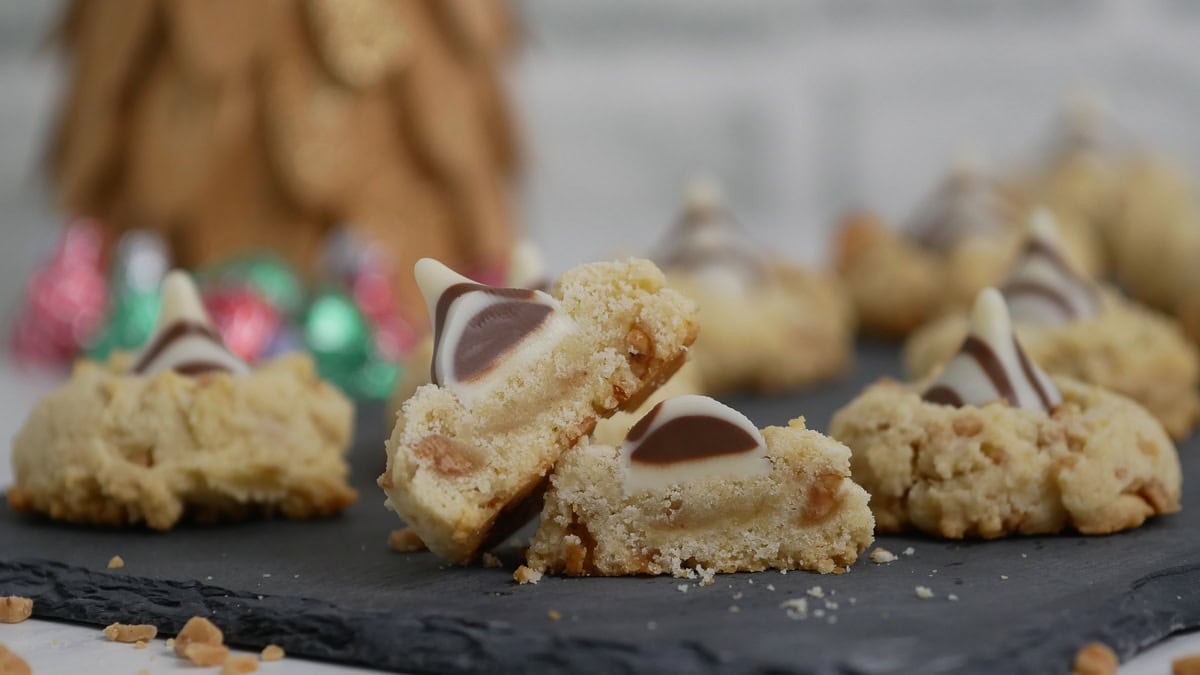 Cookies topped with striped chocolate kisses are displayed on a slate surface, with one cookie split in half to show its interior. A blurred decorative item is in the background.