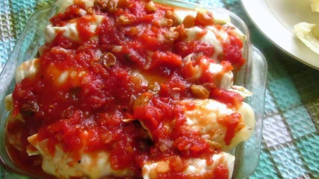 A glass dish with stuffed cabbage rolls topped with a chunky tomato sauce and raisins.