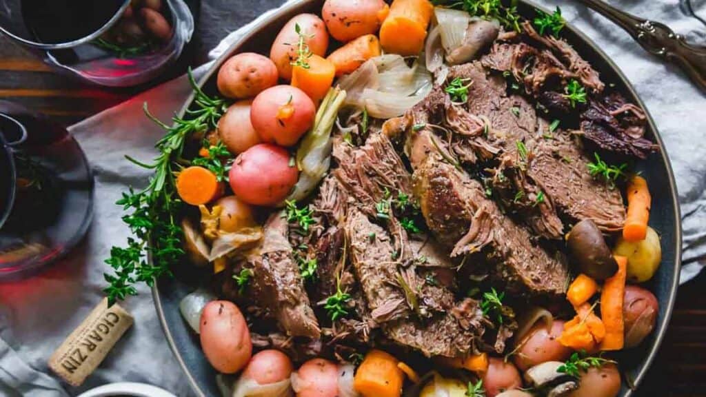 A platter of pot roast with sliced beef, carrots, potatoes, and onions garnished with fresh herbs.