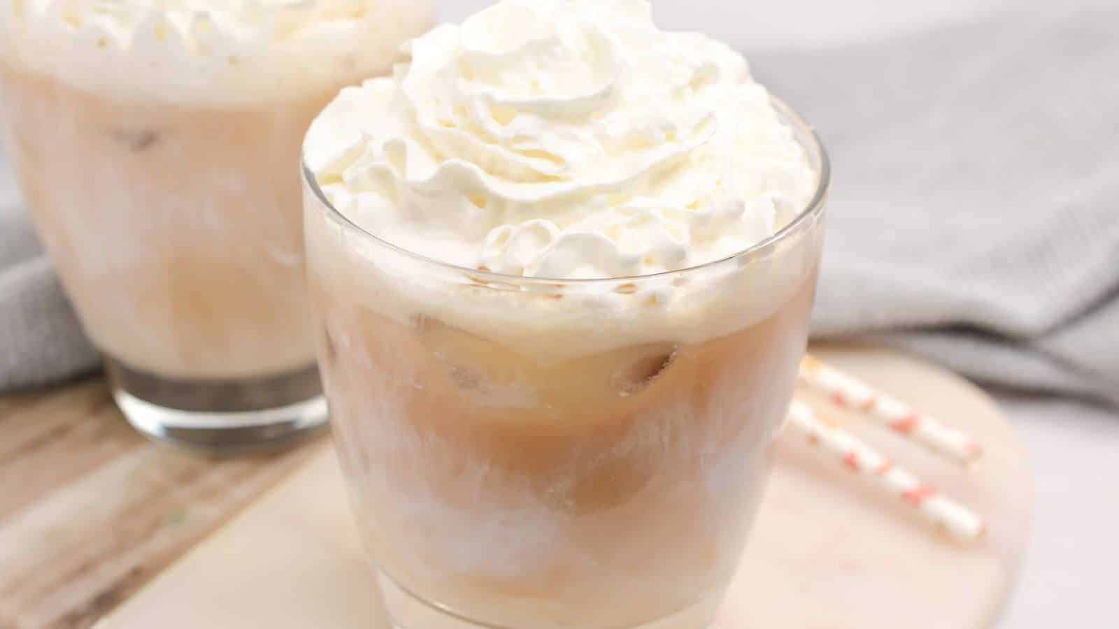 Two glasses of iced white chocolate mocha, topped with whipped cream, are on a table next to striped straws.