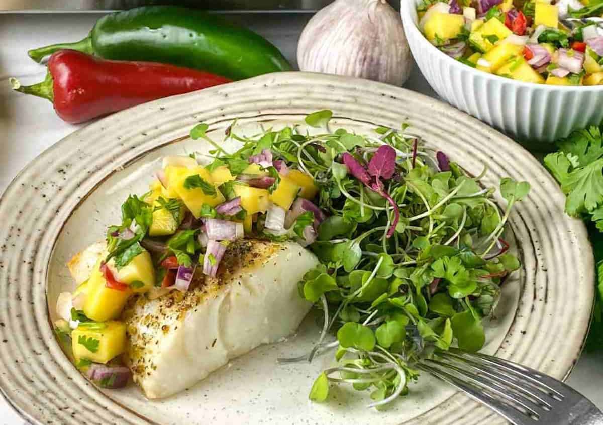 Grilled fish topped with mango salsa, served with mixed greens on a ceramic plate next to colorful peppers and a bowl of salsa.