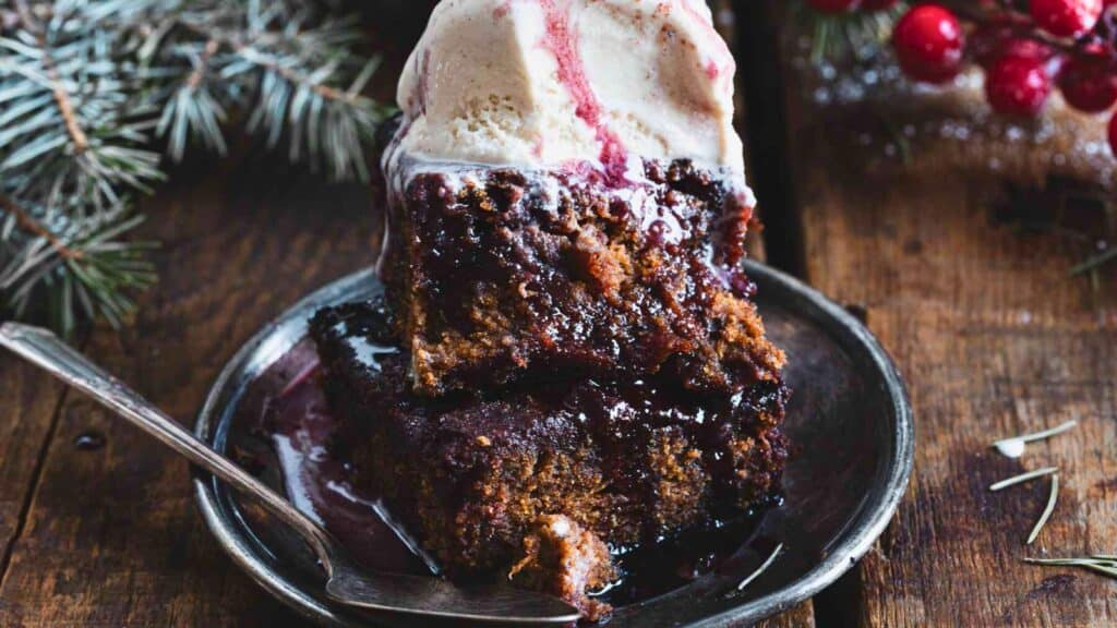 A plate of dark, moist cake topped with a scoop of vanilla ice cream and red sauce, set on a rustic wooden table with evergreen branches nearby.